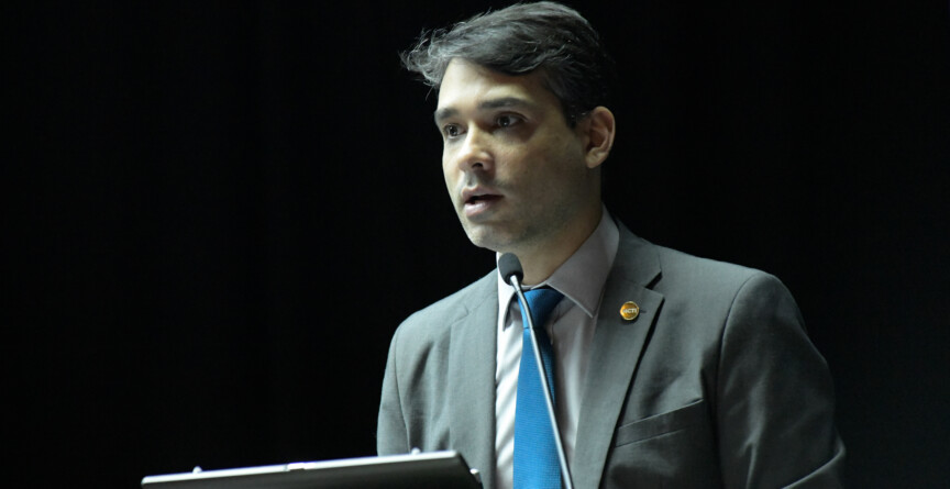 Hugo Valadares, de terno cinza e gravata azul, fala ao microfone