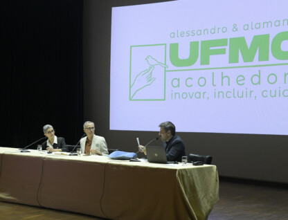 Na foto, mesa composta por Alamandra Kfouriy e Alessandro Fernandes e Gustavo Côrtes, que fala ao microfone. AO fundo, slide apresenta o slogan da chapa UFMG Acolhedora: inovar, incluir e cuidar.