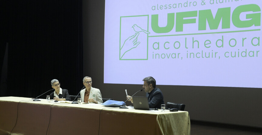 Na foto, mesa composta por Alamandra Kfouriy e Alessandro Fernandes e Gustavo Côrtes, que fala ao microfone. AO fundo, slide apresenta o slogan da chapa UFMG Acolhedora: inovar, incluir e cuidar.