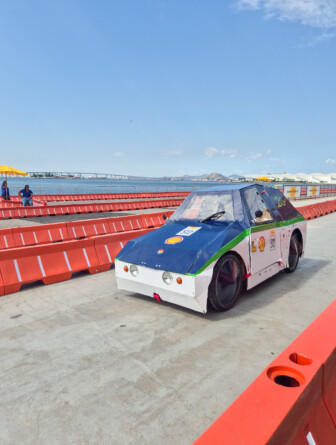 Carro azul e branco com uma listra verde ao meio em uma pista cercada por barreiras vermelhas