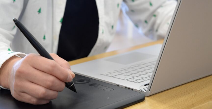 Diante de um notebook, pessoa com blusa branca faz anotações em um tablet