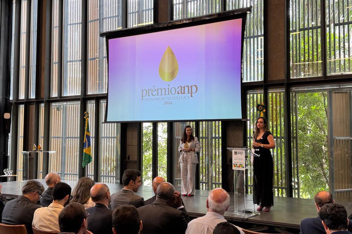 A imagem mostra uma cerimônia de premiação em um auditório com uma grande tela de projeção ao fundo. Na tela, o logotipo "prêmioanp" está em destaque com a frase "DE INOVAÇÃO TECNOLÓGICA 2024" abaixo, e uma forma dourada de gota acima. No palco, duas mulheres estão em pé. A mulher à direita, vestindo um vestido preto sem mangas, está falando em um púlpito. A mulher ao centro do palco, com roupa clara, segura um prêmio em suas mãos. Um público vestido formalmente está sentado nas cadeiras em frente ao palco. À esquerda, uma bandeira do Brasil é visível, e a arquitetura do local inclui grandes janelas com elementos verticais.