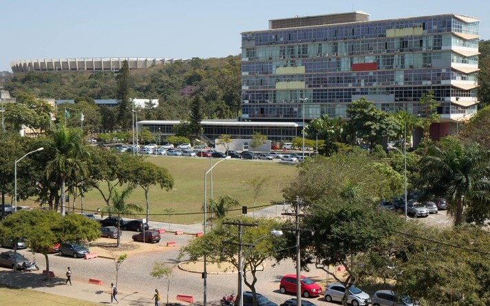 Imagem mostra o prédio da Reitoria, tendo à frente uma área verde e trecho da principal avenida do campus Pampulha, a Mendes Pimentel